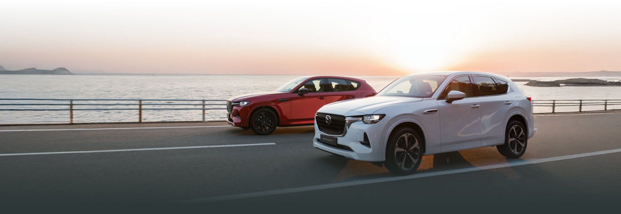 Two Mazda CX-60s driving along the waterfront with the sun setting