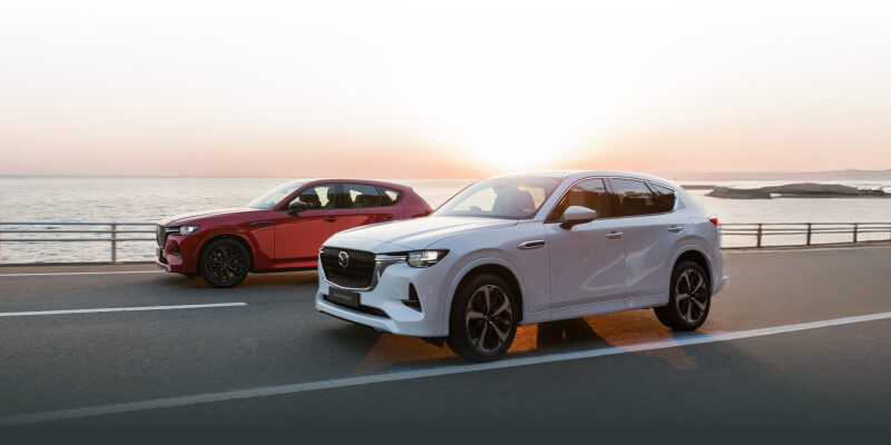 Two Mazda CX-60s driving along the waterfront with the sun setting