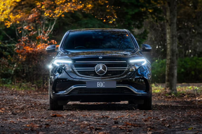 Black Mercedes-Benz EQC facing the front with headlights on and trees in the background